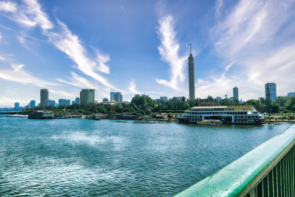 A wide-angle view of a city skyline across a river on a bright, sunny day. Tall modern buildings, including a prominent high-rise tower, are visible behind green trees lining the embankment. In the foreground, the edge of a pier or dock can be seen, with a large paddle steamer boat moored along the waterfront. The river appears calm with gentle ripples, and a smaller boat is sailing near the steamer. The sky is mostly clear with a few wispy clouds, highlighting clear weather suitable for home relocation and furniture transport activities. This scene, associated with urban moving logistics, reflects the environment where Man with Van Sipson’s removals services might operate, especially when coordinating furniture transport across city locations, including packing, loading, and unloading processes during house moves.