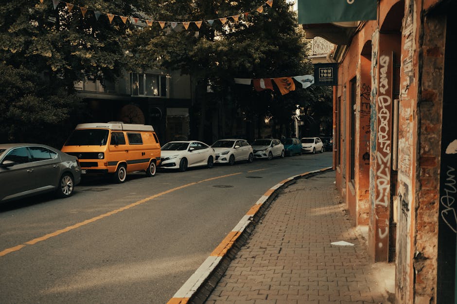 A street scene showing parked vehicles along a curbside on the left, including a grey sedan, a yellow van with a white roof, and several white cars behind. The sidewalk on the right is paved with bricks and runs beside a building with orange brick walls covered in graffiti and signage. The street appears to be part of a residential or commercial area with trees overhead, providing shade. In the background, additional parked cars and nearby buildings are visible. Occasionally, the scene suggests the context of packing and moving, as part of home relocation or furniture transport services that would be typical for house removals, with vehicles being prepared for loading or unloading. The setting is illuminated by natural daylight, capturing an everyday urban environment consistent with local removals and moving logistics, as operated by companies like Man with Van Sipson.