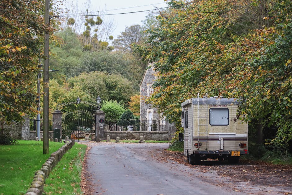 A white caravan with a ladder mounted on the rear is parked on the side of a narrow residential road bordered by green grass and low stone borders, within a leafy suburban area. The scene includes trees with autumn foliage, some with green leaves and others turning yellow and orange. In the background, there is a stone wall and a metal gate leading to a property, possibly a house or cottage, with a stone construction visible beyond the gates. The road is paved with asphalt and has some fallen leaves scattered across it, suggesting a calm, quiet environment. Utility poles and wires run along the left side of the street, indicating electrical infrastructure. This setting captures elements typical of a home relocation or moving process, with the caravan positioned to support furniture transport, packing, and loading activities. The environment appears well-maintained, with soft natural lighting suggesting a daytime scene, complementing the services offered by Man with Van Sipson for local removals and relocation needs.