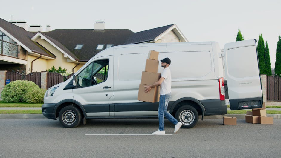 A man wearing a dark jacket, light-colored sneakers, and a cap is standing at the back of a white delivery van with open Rear doors, surrounded by a paved outdoor area. Inside the van, there are stack