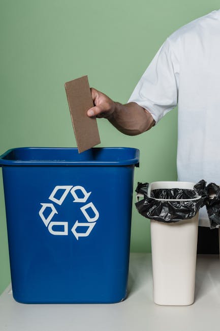 A person wearing a white shirt is holding a rectangular cardboard piece above a large blue recycling bin with a white recycling symbol on it. Next to the blue bin, there is a smaller white bin lined with a black plastic bag, which is partially open and filled with waste items. The scene takes place indoors, with a plain green wall in the background. This setup reflects waste disposal during home relocation or packing processes, with bins positioned on a flat surface, ready for sorting and recycling materials before moving. The image emphasizes the careful sorting of waste, which is an integral part of house removals and furniture transport services offered by Man with Van Sipson, helping facilitate smooth and environmentally conscious moves.