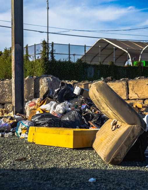 An outdoor scene showing a pile of bulky waste and household clutter consisting of black and white plastic rubbish bags, an overturned old beige upholstered armchair, and a yellow plastic storage container placed on a gravel surface near a stone wall. The background includes a metal fence, a green hedge, utility poles with overhead wires, and a partly cloudy blue sky. The image appears to be taken during daylight hours, possibly during a waste clearance or property cleanup as part of a house removal process. The clutter and furniture are positioned on the pavement outside a building, with no people visible. This scene may relate to the disposal of unwanted items before a home relocation, consistent with the services offered by Man with Van Sipson in the removals category.