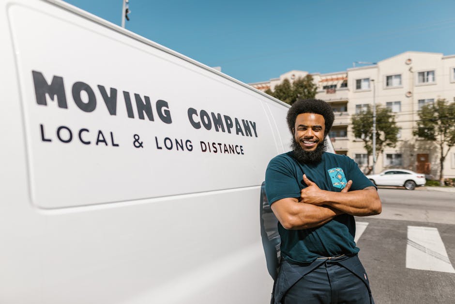 Two movers from Man with Van Sipson are seated inside the open rear of a white moving van parked on a street. They are wearing casual work uniforms with dark pants and long-sleeved shirts, smiling at