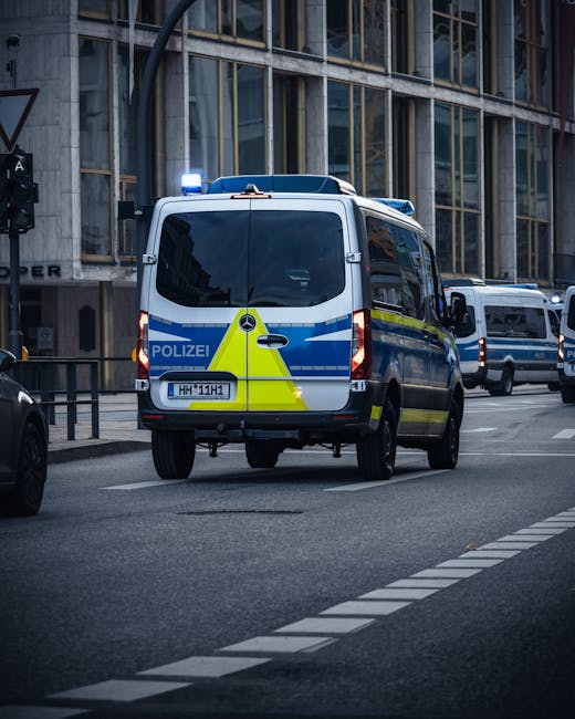 A police van with a blue and yellow chevron pattern and flashing blue lights on top is parked on a city street in front of a modern building with large glass windows. Several other vehicles are visibl