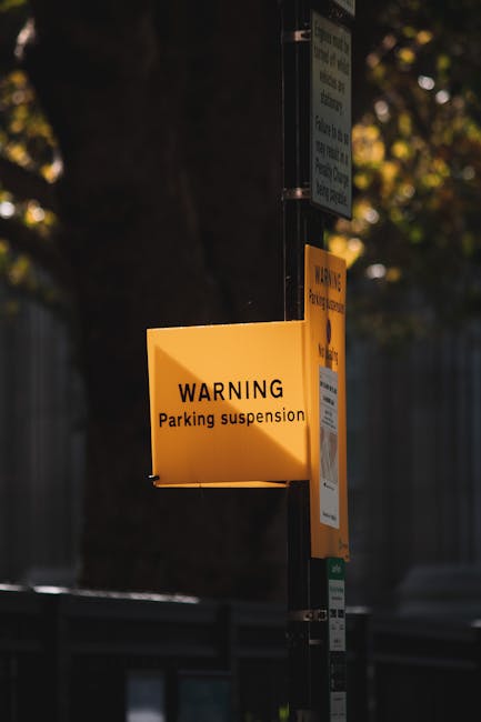 A bright yellow rectangular sign attached to a metal pole displaying a warning about parking suspension, with the text 'WARNING Parking suspension' clearly visible. The sign is outdoors, with blurred dark background elements including trees and fencing, and is illuminated by natural light. The sign is situated near a residential or commercial area, indicating restricted parking during specific times, which could affect house removals or moving services. This visual relates to parking regulations in Sipson, as referenced on the page about Hillingdon Council's parking suspensions and fines, and highlights the importance of coordinating parking permits and suspensions during home relocation or furniture transport operations managed by Man with Van Sipson.