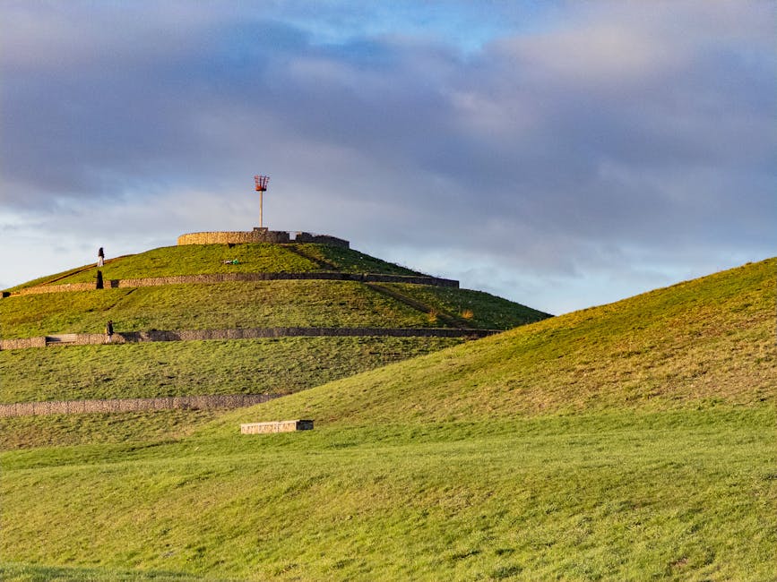 A large grassy hill with terraced levels and stone retaining walls, leading up to a circular stone platform at the summit with an orange and white navigation tower. The hill is illuminated by daylight, casting shadows on the slope, with a partly cloudy sky overhead. There are a few small groups of people visible on different levels of the terraced grass, and the area appears to be a public outdoor space. The image captures the landscape in the context of an urban park or historic site, which may relate to local transportation or outdoor activities, as referenced in the council parking suspensions and removals services offered by Man with Van Sipson.