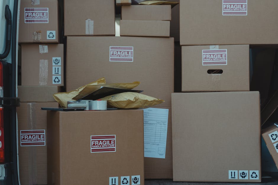 A collection of packed cardboard boxes of various sizes, some labeled with 'FRAGILE Handle with Care' stickers, is shown loaded into the back of a van. The boxes are arranged on the vehicle's floor, with a few partially covered by brown packing paper and a plastic-wrapped item. The scene depicts a home relocation process, with boxes awaiting transport, inside a vehicle designed for removals. The environment appears to be well-lit, highlighting the neatly stacked boxes and packing materials, which are typical for furniture transport and moving services. Several icons indicating proper handling and recycling are visible on the boxes, emphasizing careful packing and eco-conscious disposal. The image illustrates the logistics involved in packing and moving large items and reflects the professional approach of [COMPANY_NAME] during a house move near Heathrow, supporting seamless relocation and furniture transport processes.
