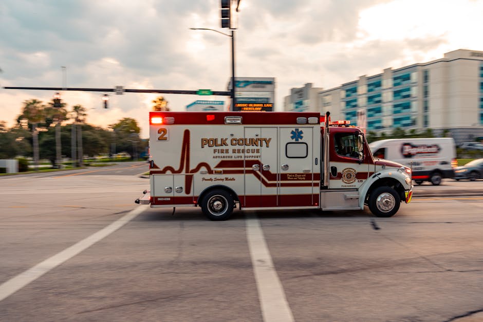 A red and white Polk County Fire Rescue ambulance vehicle parked on an urban street at dusk, with a partially cloudy sky overhead. The ambulance has emergency lights on top, a side door, and the Polk County Fire Rescue logo. In the background, there are multi-storey residential or commercial buildings, traffic lights, and other vehicles, including a white van. The scene shows the vehicle in motion, with a slight blur indicating movement, and the surrounding environment includes road markings and street infrastructure. This image relates to emergency transportation as part of logistical support for home relocations or urgent services, reflecting professional vehicle use aligned with house removals and moving logistics.