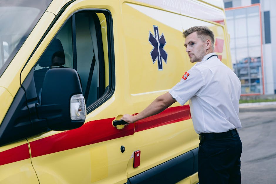 A male emergency medical technician with light skin and short, styled blond hair is standing beside a yellow ambulance with red reflective stripes and a blue medical cross symbol on the side. He is wearing a white medical uniform shirt with a red cross patch on the sleeve and dark trousers. The technician is holding the ambulance door handle with his right hand, preparing to open the door, and is facing slightly to the right with a focused expression. The scene is outdoors on a paved area, with a modern glass building and a partly cloudy sky in the background. The ambulance's side mirror, window, and part of the vehicle's body are visible, along with a red emergency fire alarm on the side of the vehicle. This setting suggests a quick response scenario involving the transport of patients in a house removal or medical emergency situation, with the vehicle ready for use in relocation or urgent medical transport. Man with Van Sipson offers services aligned with emergency and same-day home relocations, as indicated on the webpage, illustrating the staging of rapid transport and logistical support in moving services.