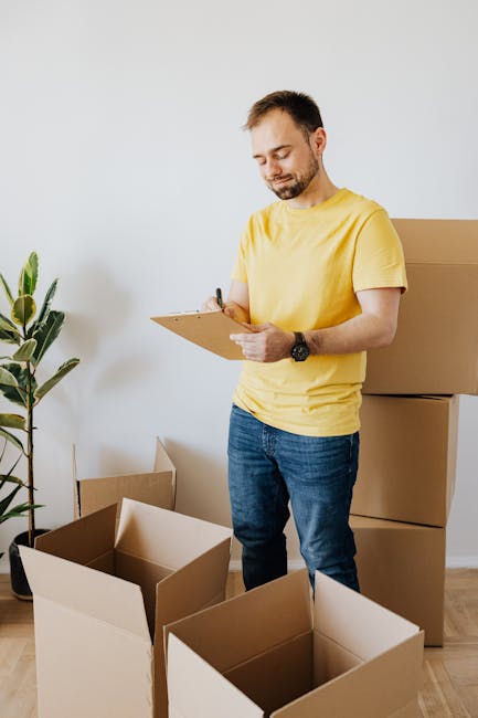 A man with a beard and short dark hair, dressed in a yellow t-shirt and blue jeans, is standing indoors next to a stack of cardboard moving boxes. He is holding a clipboard and a pen, appearing to review or check a list related to house relocation or packing. Behind him, there are additional boxes arranged against a plain white wall, with some partially assembled or open, ready for packing or transportation. To his left, a tall green potted plant adds natural decor to the space. The setting suggests an organized packing or furniture transport process as part of a home removal or move, which is typical for services provided by Man with Van Sipson. The lighting is natural, and the environment looks tidy and well-prepared for a moving day, reflecting the preparation stage of packing and moving logistics, as outlined in the Sipson Lane Apartment Move Checklist.