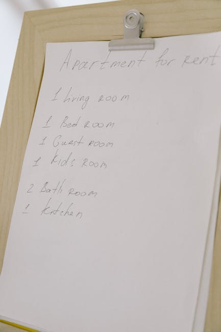 A close-up photograph of a handwritten apartment move checklist taped to a wooden surface with a white clip. The list, written in pencil or pen, details various rooms such as the living room, bedroom, guest room, kids' room, bathrooms, and kitchen, indicating areas involved in a home relocation. The paper is slightly curled at the edges, and the background suggests an indoor setting with soft, natural lighting. This checklist is relevant to house removals and packing preparations. Man with Van Sipson, a professional removal service, might use such a list during the furniture transport and moving process to ensure proper organization when relocating belongings between properties.