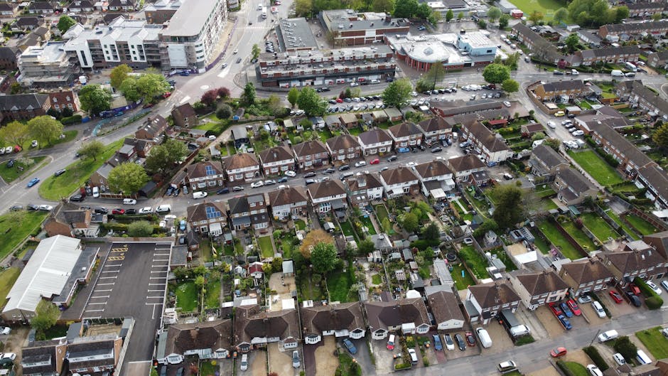 An aerial view of a residential neighbourhood with rows of terraced houses featuring small back gardens, some of which have outdoor furniture and garden sheds. A few houses are equipped with solar panels on their roofs. The street in front of the houses is lined with parked cars, and the houses are separated by narrow pathways and fences. To the left, there is a large, flat-roofed commercial building with a spacious parking lot nearby. In the background, a busy urban area with multi-storey buildings, commercial properties, and a parking area can be seen, along with green trees and open spaces. This scene depicts a typical suburban area suited for home relocation and furniture transport services. Man with Van Sipson can assist with packing, loading, and transporting household items through this neighbourhood as part of their house removals and moving logistics, ensuring a smooth collection and delivery process during a home move or relocation project.