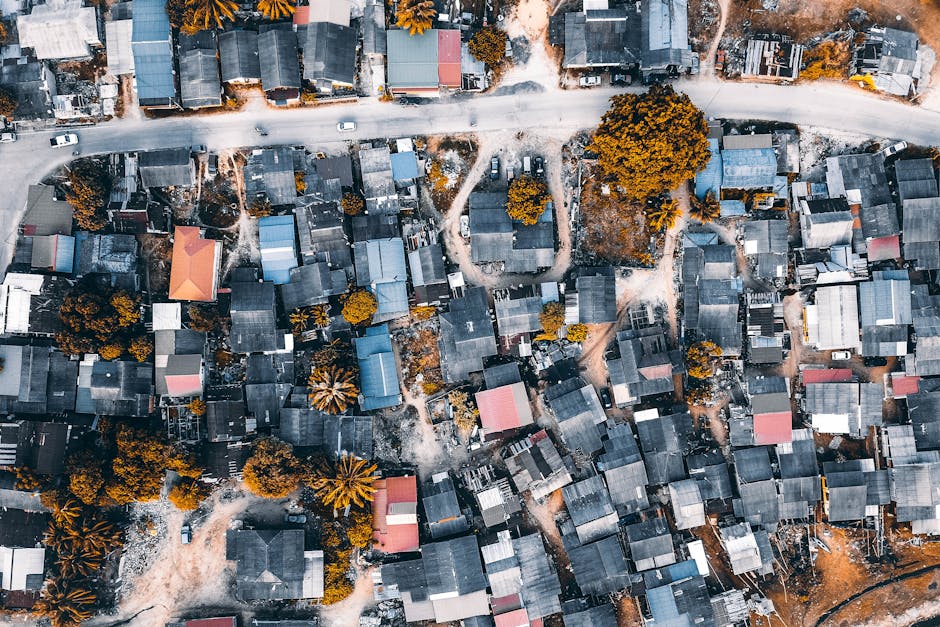 An aerial aerial view of a densely packed residential area showing numerous small houses with flat and gable roofs in shades of gray, black, and brown, interspersed with a few trees exhibiting autumn foliage in shades of orange and brown. The houses are arranged along narrow, winding pathways and alleys, with some feet and a few vehicles visible on the roads. The buildings are closely clustered together, indicating a compact neighbourhood. The image captures the overall layout and urban density, which may be relevant for planning house removals and furniture transportation in Sipson Village, with some parts of the area appearing to be undergoing ongoing work or renovation, as suggested by the presence of construction materials or equipment visible around certain structures.