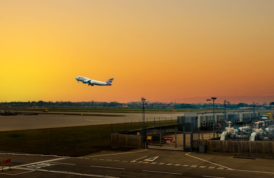 A British Airways commercial airplane is mid-flight, ascending from Heathrow Airport during sunset, with a vibrant orange and yellow sky in the background. The aircraft's fuselage and tail fin with the airline's logo are clearly visible. In the foreground, the airport tarmac displays various ground equipment, including fuel tanks, pipelines, and service structures, all enclosed within a secure perimeter fence. The scene captures part of the airport's operational area with control towers and additional runway infrastructure. This scene reflects the busy logistics involved in international air travel and home relocation services, relevant to moving and packing processes managed by companies like Man with Van Sipson, especially in the context of adhering to move rules around Heathrow, licenses, and safety regulations.