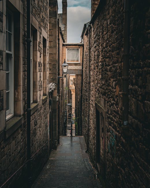 A narrow cobbled street between two tall, historic brick buildings with small windows and black drainpipes, illuminated by a vintage-style street lamp attached to the left building. The street is slightly wet, with a gentle slope downward, leading to a view of more buildings and a cloudy sky in the distance. No vehicles or furniture are visible in the scene. The setting suggests an old urban area suitable for house removals in tight streets, where careful planning and possibly special permit advice, as outlined in the UB7 Removals Guide by Man with Van Sipson, is necessary for home relocation services involving furniture transport and loading within confined alleyways.