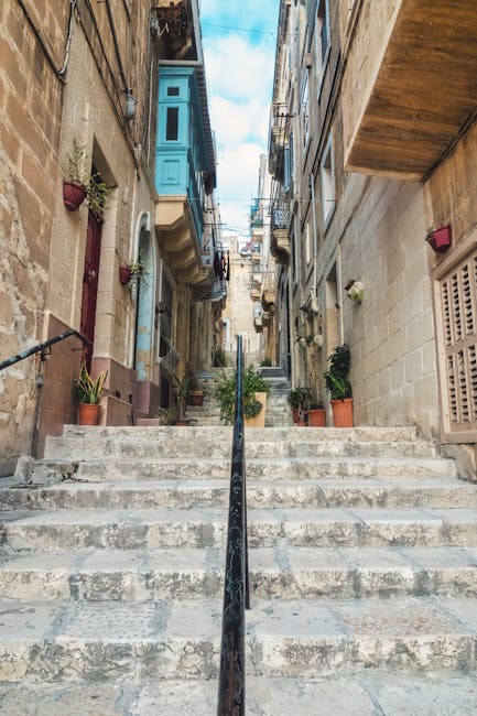 A view of a narrow outdoor stairway in a residential area, with stone steps leading up between historic buildings on either side. The staircase has a black metal handrail running along the centre, and potted plants are placed along the edges of the steps. On the left, there is a building with a stone facade, featuring small windows and flower boxes, while on the right, a building with a plain wall and a beige vent covers part of its exterior. Above, the sky is partly cloudy, and the scene suggests a quiet, community-oriented environment. This setting is typical of urban home relocations, where careful handling of furniture and packing materials is necessary for moving within narrow streets, as managed by specialists like Man with Van Sipson, ensuring safe and efficient delivery of items during the moving process.