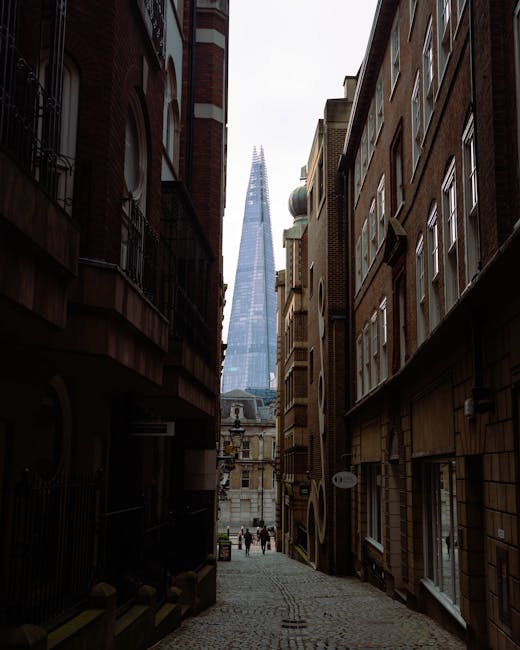 A narrow cobbled street between two tall, historic brick buildings with small windows and black drainpipes, illuminated by a vintage-style street lamp attached to the left building. The street is slightly wet, with a gentle slope downward, leading to a view of more buildings and a cloudy sky in the distance. No vehicles or furniture are visible in the scene. The setting suggests an old urban area suitable for house removals in tight streets, where careful planning and possibly special permit advice, as outlined in the UB7 Removals Guide by Man with Van Sipson, is necessary for home relocation services involving furniture transport and loading within confined alleyways.